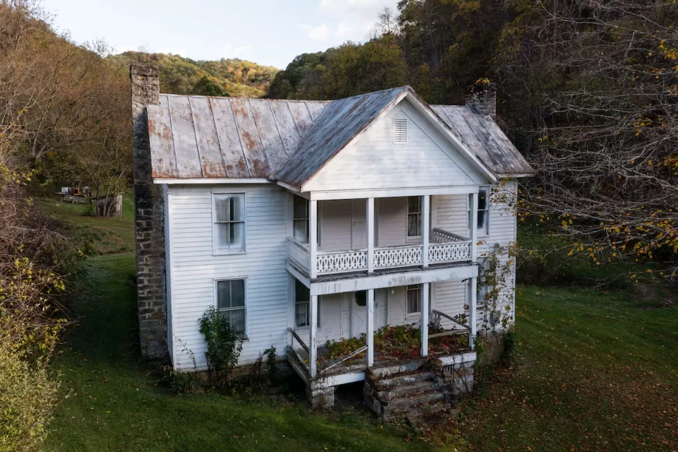 White two-story abandoned farmhouse in lush green field surrounded by trees in West Virginia.